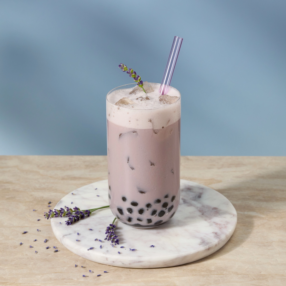 A tall glass of iced bubble tea with black tapio pearls at the bottom and a purple-striped straw, set on a round white marble coaster with lavender sprigs on a light wood surface against a soft blue background.
