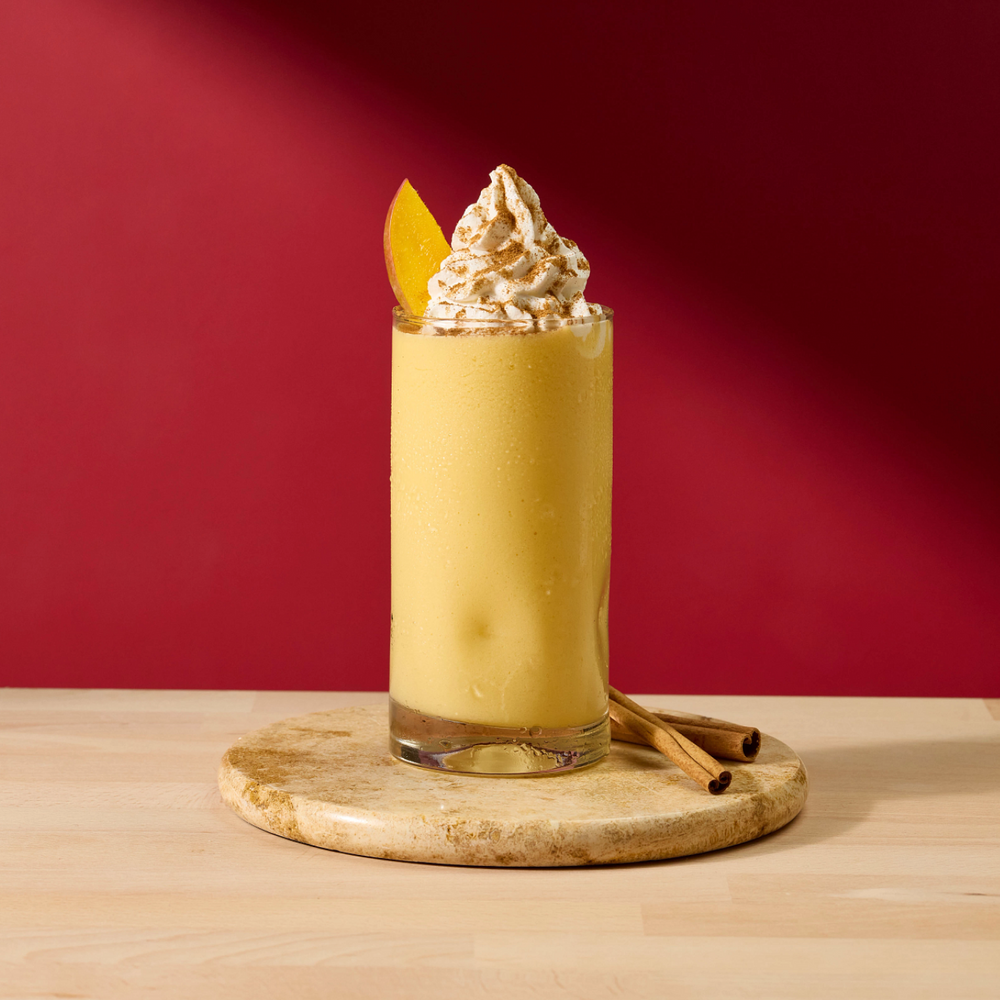 A tall glass filled with a yellow-orange beverage, topped with whipped cream and garnished with a citrus slice, set on a round wooden coaster with cinnamon sticks against a deep red background.