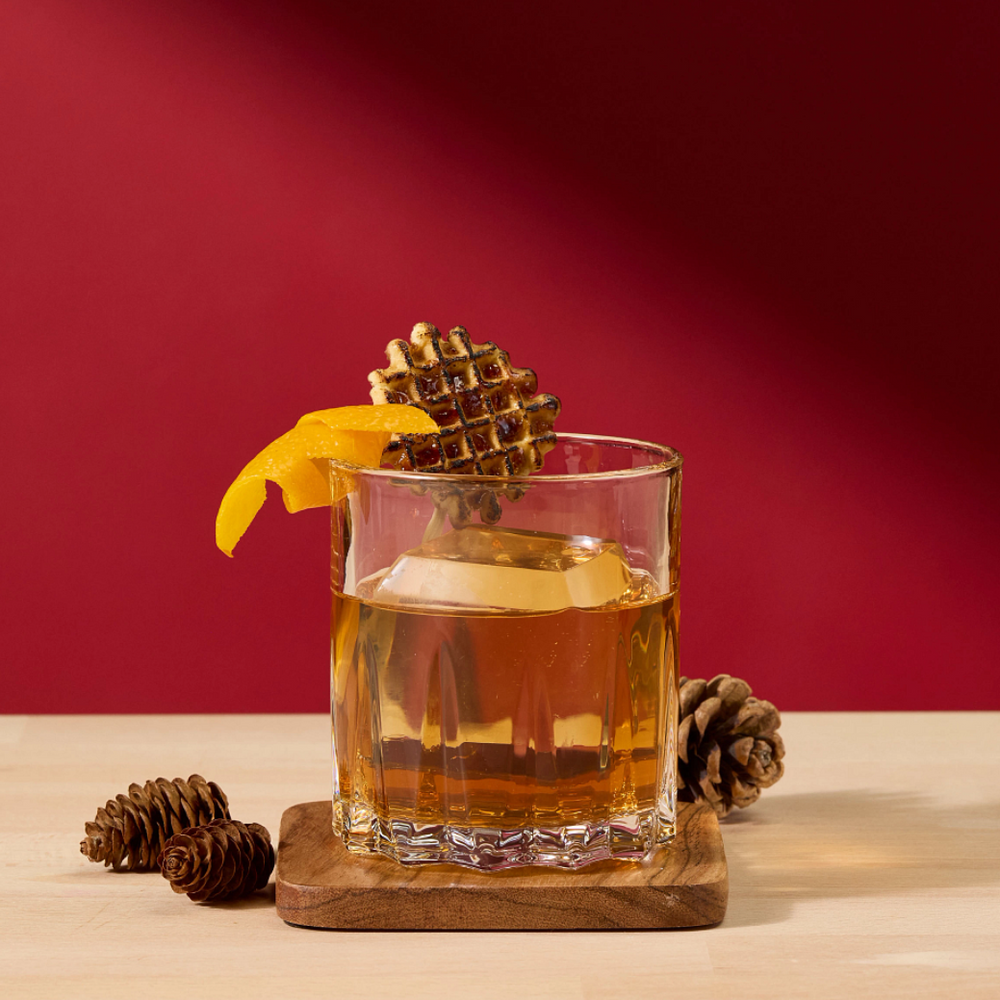 A clear rocks glass filled with an amber drink, garnished with a orange peel twist and a pinecone, set on a wooden coaster on a light wood surface against a dark red background.