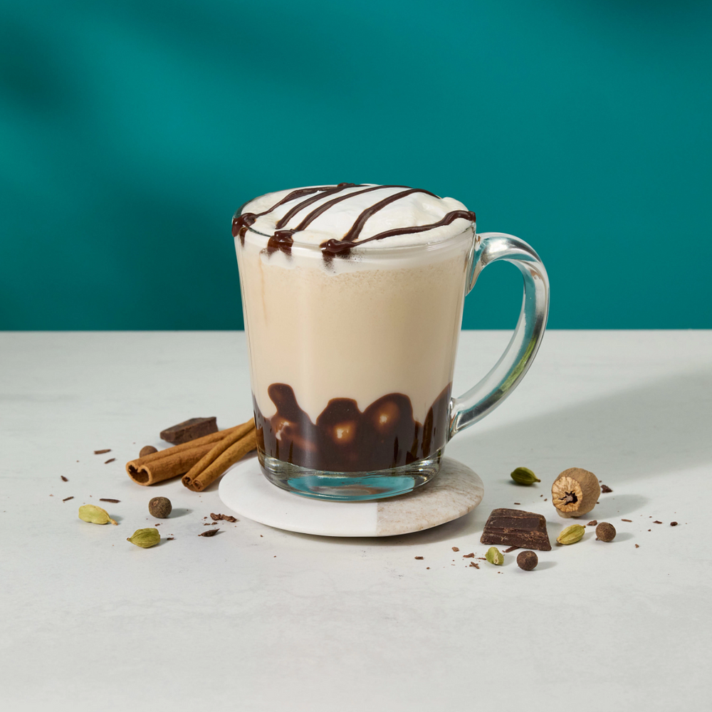 A frothy latte in a clear glass mug with a handle, topped with chocolate drizzle, set on a white coaster with cinnamon sticks, nuts, and chocolate pieces scattered around it on a white surface against a teal background.