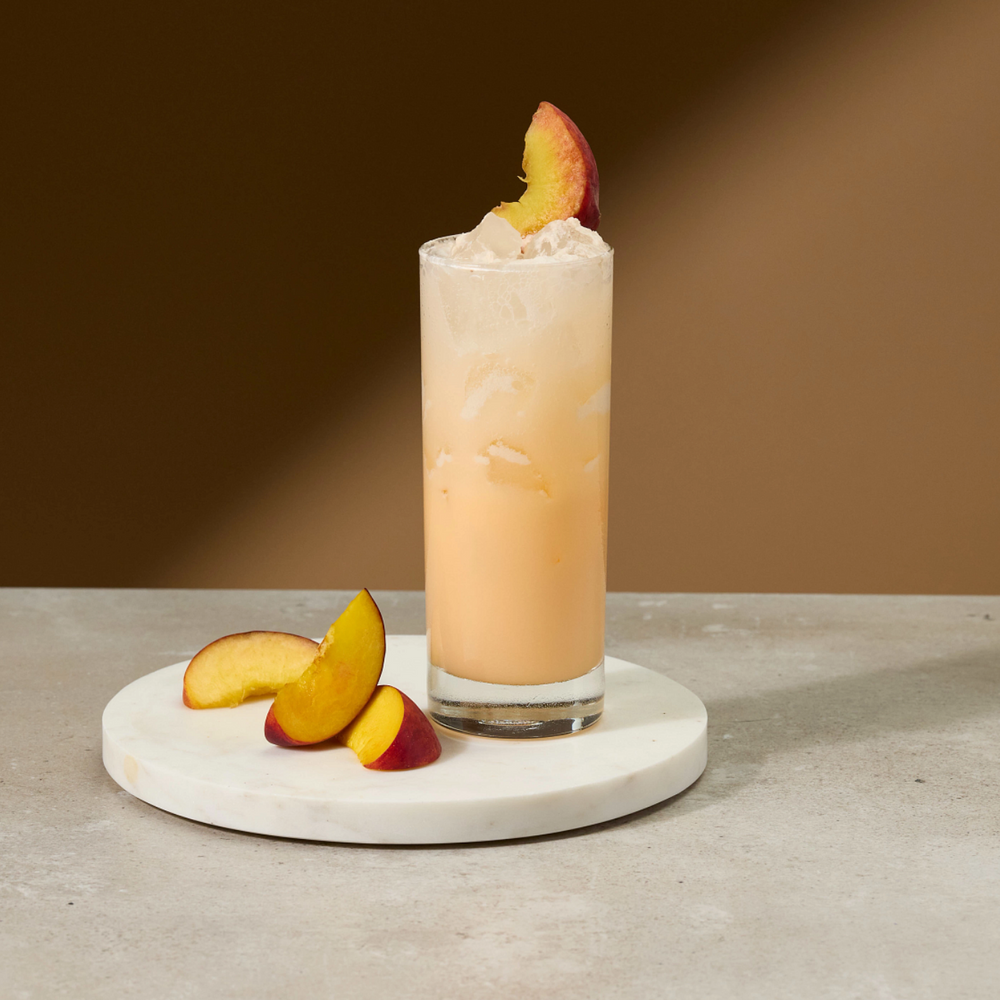 A tall clear glass filled with an orange-amber beverage and topped with whipped cream and a peach slice, set on a round white coaster with peach slices on a light stone surface against a warm brown background.