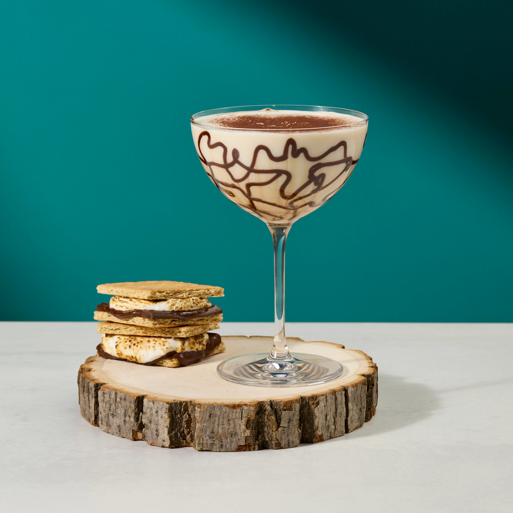A dessert coupe glass filled with a light cream or chocolate mousse, topped with dark chocolate drizzle, displayed on a round wood slice coaster next to a small stack of golden-brown cookies against a teal background.
